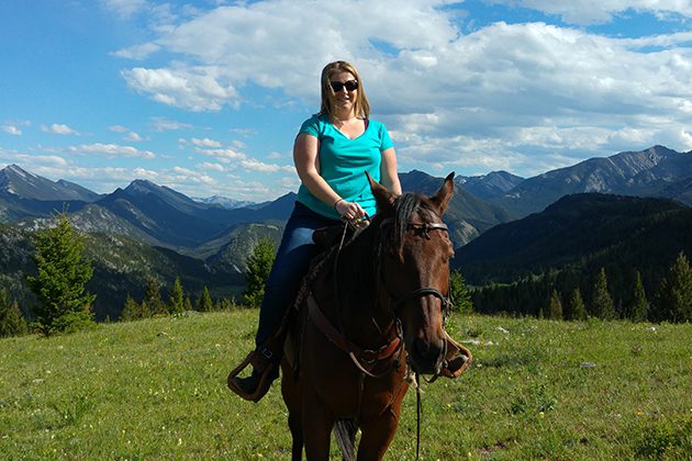 A woman riding on the back of a brown horse among greenery and mountains