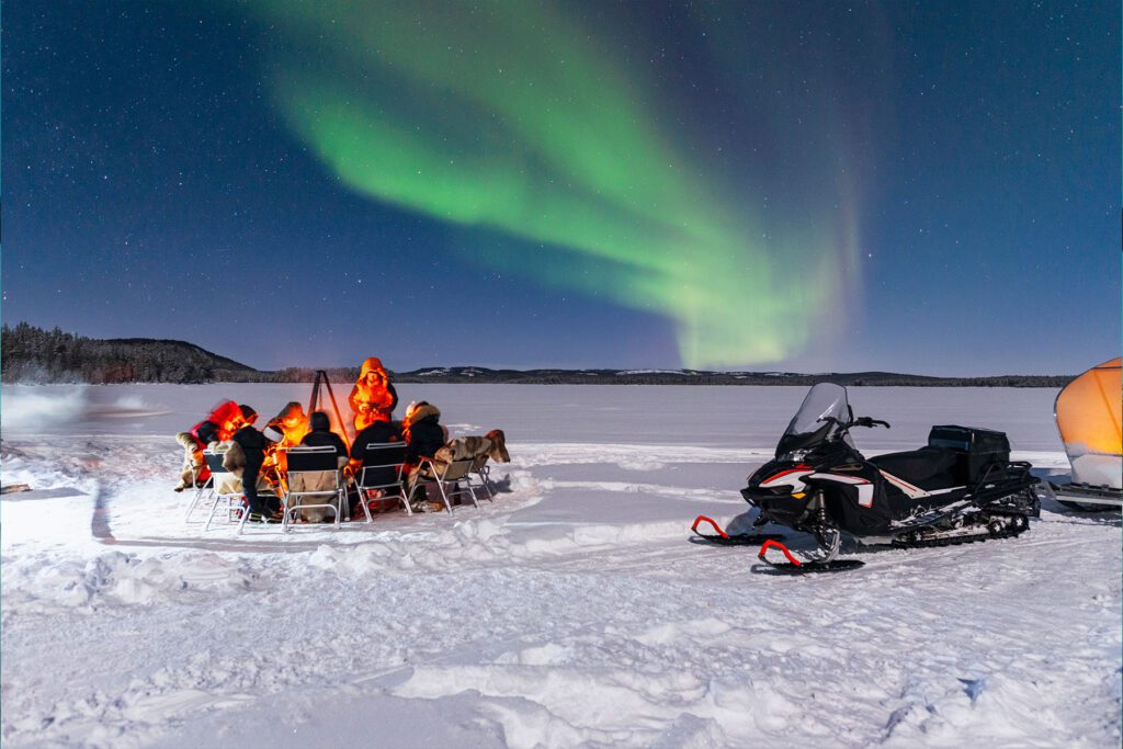 people huddled around a fire with the northern lights in the background and a snowmobile in the foreground