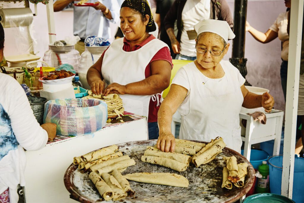 Medium wide shot of female workers cooking traditional Oaxaca street food at outdoor market stall during lunch time rush