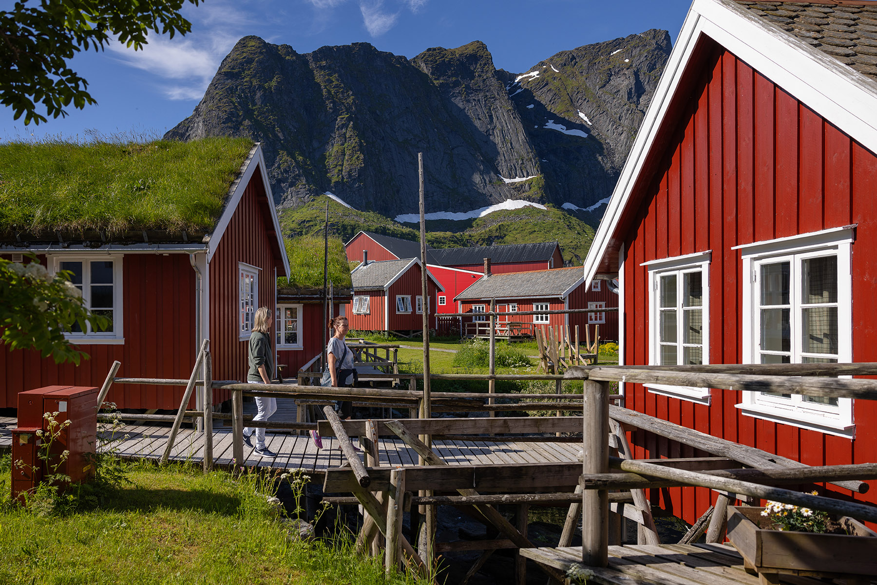 two people walking across a bridge inbetween red houses with grass growing on the roof
