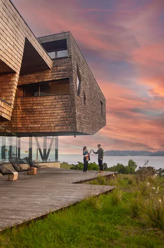 People drinking wine in front of a lodge hotel with a lake and mountains in the background